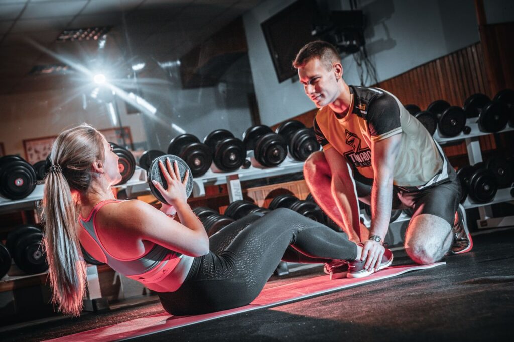 A personal trainer assists a woman doing sit-ups with a medicine ball in a gym setting.
