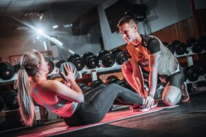 A personal trainer assists a woman doing sit-ups with a medicine ball in a gym setting.